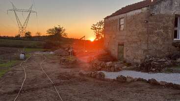 A rustic stone house stands beside a gravel path at sunset, with power lines in the distance and trees silhouetted against the orange glowing sky.