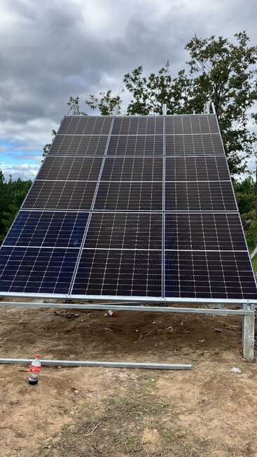 This image shows a large solar panel installation outdoors on a metal frame, with cloudy skies above, trees in the background, and a soda bottle on the ground.