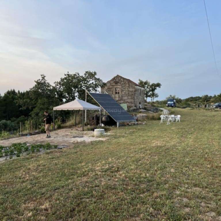 A rustic scene shows an old stone building, solar panels, white canopy, outdoor chairs, a person walking, greenery, grassy field, parked car, and trees under blue sky.