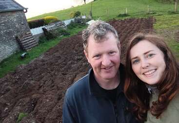The image shows a man and woman standing in front of a plowed field, with a stone building and green hills in the background.