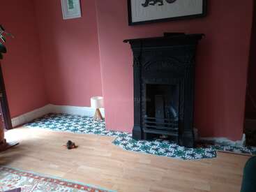 A cozy room with pink walls features a black ornate fireplace, patterned tiles, a wooden floor, a small lamp, artwork, and a toy bird on the floor.