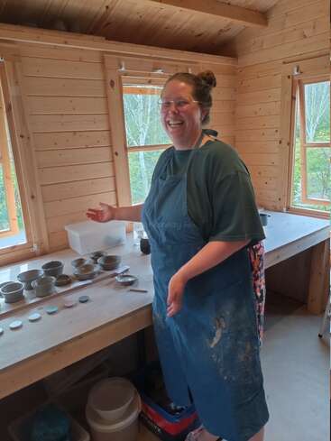 A smiling woman in glasses and an apron stands in a wooden studio, presenting handmade pottery on a table. The room has large windows and natural light.