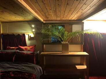 A cozy room with wooden ceilings, a comfortable bed with red pillows, a wooden table, a large green plant, and warm, ambient lighting.