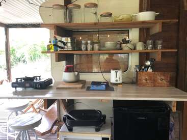 This image shows a rustic kitchen setup with open wooden shelves holding jars, dishes, and glasses. The counter has appliances, utensils, and a small fridge.