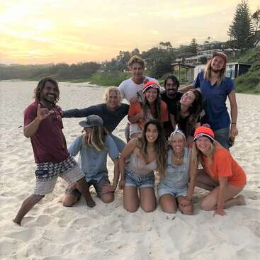 La imagen muestra a un grupo de 11 personas posando en una playa de arena, con casas y árboles visibles al fondo al atardecer.