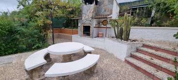 This image shows an outdoor patio with a stone barbecue grill, round stone table with benches, cactus plants, garden greenery, and red-tiled steps leading upward.