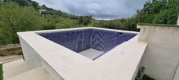 This image shows an empty, square, outdoor pool with blue tiles. It is surrounded by greenery and located on a hillside under a cloudy sky.