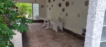 This image shows a covered outdoor patio with a white table, stacked white chairs, wall-mounted ceramic plates, textured floor, green plants, and natural light.
