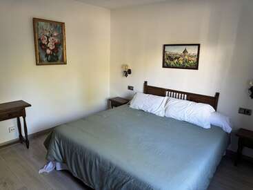 This image shows a cozy bedroom with a large bed, wooden headboard, two bedside tables, wall-mounted lamps, and two paintings on pale walls. Neatly arranged.