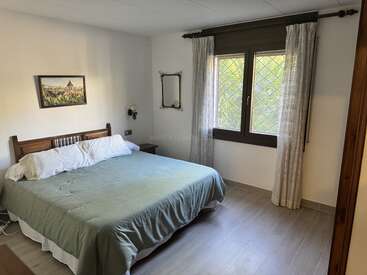 This cozy bedroom features a double bed with green bedding, wooden headboard, wall art, a mirror, bedside table, large window with curtains, and tiled floor.
