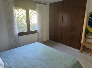 This bedroom features a large window with sheer curtains, a simple bed, wooden wardrobe, tiled floor, a chair, and a partial view of a flag.