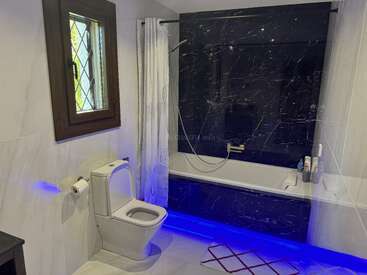 This modern bathroom features a toilet, window, bathtub with black marble walls, white shower curtain, and striking blue LED lighting underneath, creating a clean, stylish ambiance.