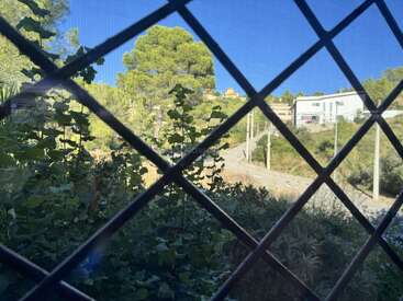 A metal grid covers the window view of lush greenery, a dirt path, electricity poles, trees, and white houses under a clear blue sky.