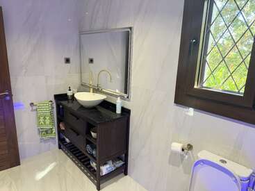 This modern bathroom features a vessel sink, black countertop, wooden vanity, large mirror, towel rack with green towel, window with grille, toilet, and marble tiles.