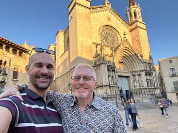 Two smiling men take a selfie in front of a grand, historic cathedral. The intricate architecture and clear blue sky create a beautiful, joyful travel moment.