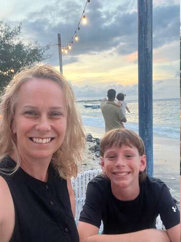 Una mujer y un niño sonríen a la cámara cerca de una playa al atardecer. Encima brillan cuerdas de luces y un hombre sostiene a un niño en la orilla.