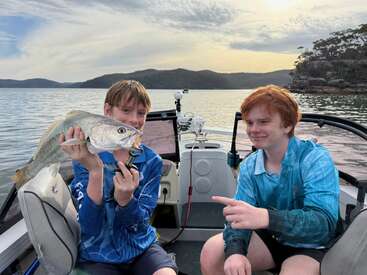 Al atardecer, dos chicos sonríen orgullosos sentados en una barca. Uno sostiene un pez recién pescado, mientras el otro lo señala, celebrando su éxito pesquero.