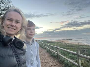 Una mujer y un niño sonríen en un camino de arena junto al océano al atardecer, con cielo nublado, hierba verde y vallas de madera al fondo.