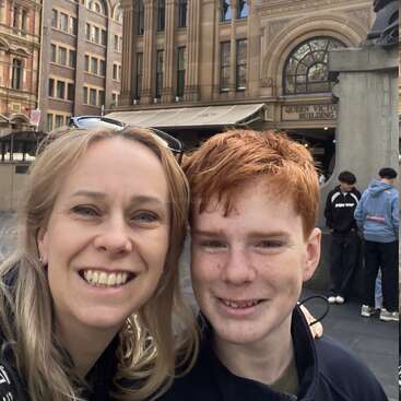 Uma mulher e um adolescente de cabelos vermelhos sorriem para uma selfie em frente ao Queen Victoria Building. Pessoas e arquitetura histórica preenchem o plano de fundo.