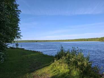 Ein ruhiger Fluss fließt sanft unter einem klaren, blauen Himmel, umgeben von saftig grünem Gras, Büschen und fernen Wäldern, die an eine friedliche, natürliche Landschaft erinnern.