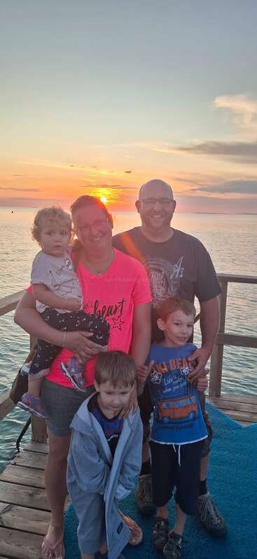 Eine fünfköpfige Familie lächelt bei Sonnenuntergang auf einem Steg und genießt einen friedlichen Moment am Wasser mit strahlendem Himmel und warmer, fröhlicher Atmosphäre.