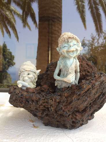 La imagen muestra dos pequeñas estatuas blancas de personas sonrientes sobre una roca marrón oscura, con palmeras y un sereno cielo azul como telón de fondo.