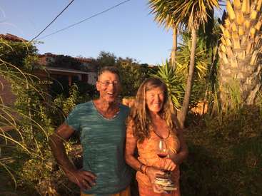 La imagen representa a un hombre y una mujer juntos en un entorno tropical, rodeados de palmeras y otra vegetación, con la mujer sosteniendo un vaso de vino.