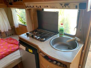 La imagen muestra el interior de una autocaravana, con una pequeña cocina con fregadero y fogones, una cama pequeña y una ventana con vistas al exterior.