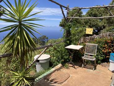 La imagen muestra un sereno patio al aire libre con una silla de madera, una pequeña mesa y una exuberante vegetación, con vistas a una vasta masa de agua bajo un cielo azul despejado.