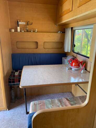 La imagen muestra el interior de una furgoneta camper, con una mesa, un banco y una ventana con vistas a los árboles del exterior, en un espacio acogedor y compacto.
