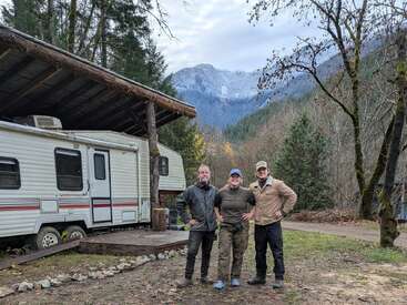 Drei Leute stehen fröhlich draußen neben einem Wohnmobil unter einem Holzunterstand, umgeben von Bäumen und mit Bergen im Hintergrund. Die Szene wirkt friedlich und rustikal.