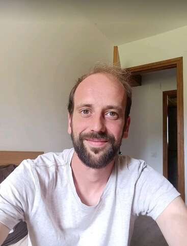 A man with a beard and short hair smiles at the camera, wearing a light gray shirt, sitting indoors with a wooden door and neutral background.