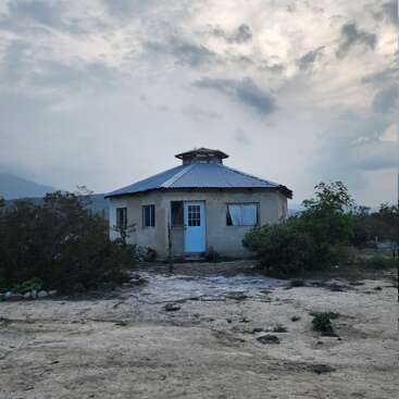 Ein kleines, rustikales achteckiges Haus mit einem Metalldach steht ganz allein in einer trockenen, halbwüstenartigen Landschaft, umgeben von vereinzelten Büschen unter einem dramatisch bewölkten Himmel.