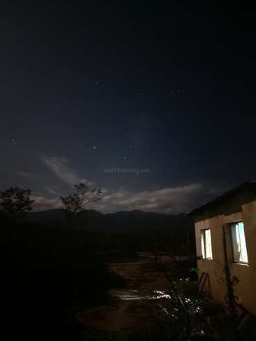 Une scène nocturne sombre et paisible montre une maison aux fenêtres éclairées, entourée de silhouettes d'arbres et de montagnes, sous un ciel clair rempli d'étoiles.