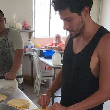 Trois personnes se trouvent dans une cuisine ; deux hommes préparent de la pâte sur un comptoir, tandis qu'une femme âgée est assise à une table à l'arrière-plan, souriante.