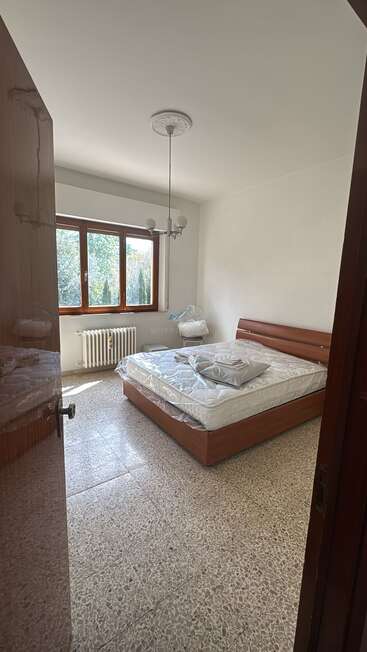 La imagen representa un dormitorio con cama, ventana y puerta, con paredes blancas, somier marrón y suelo de terrazo, y una vista de árboles al otro lado de la ventana.