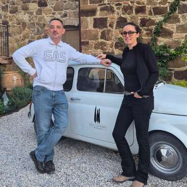 La imagen muestra a un hombre y una mujer de pie junto a un pequeño coche blanco, con un edificio de piedra como telón de fondo. La pareja viste un atuendo informal.
