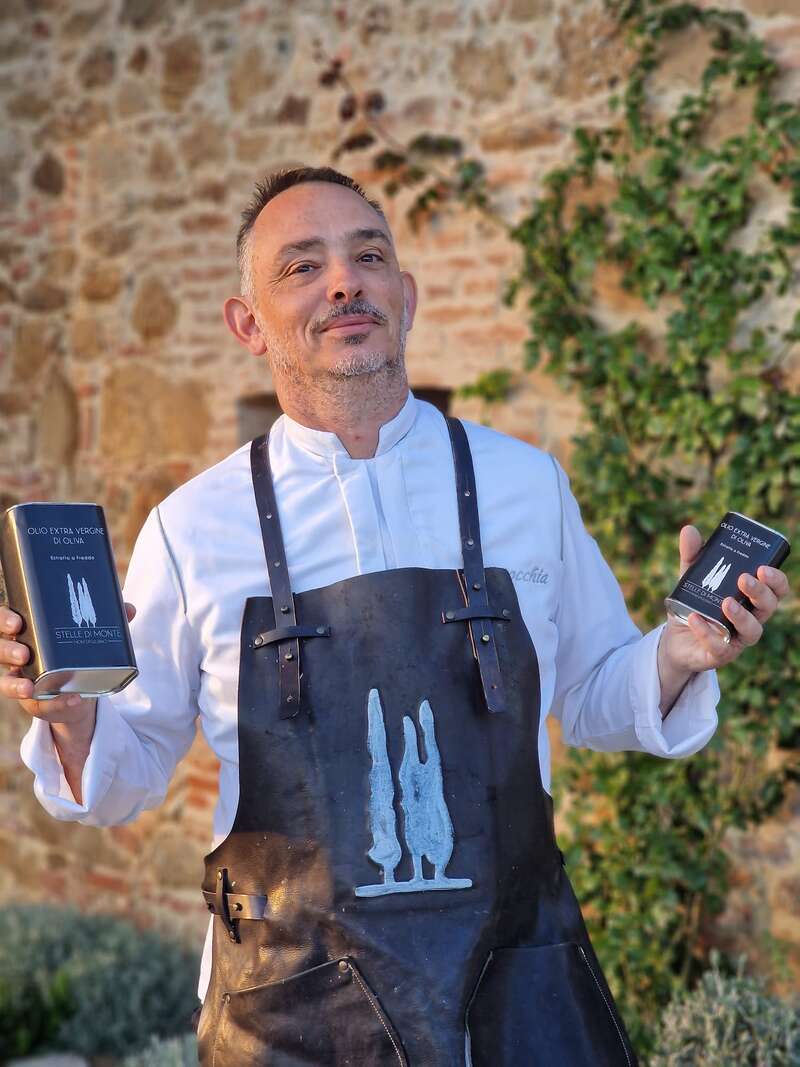 La imagen muestra a un hombre con camisa blanca y delantal negro, sosteniendo dos latas de aceite de oliva, de pie frente a un muro de piedra con vegetación.