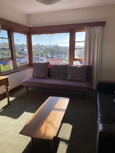 L'image représente un salon confortable avec une grande fenêtre, un canapé et une table basse, baigné par la lumière du soleil, avec une vue sereine sur le quartier environnant.