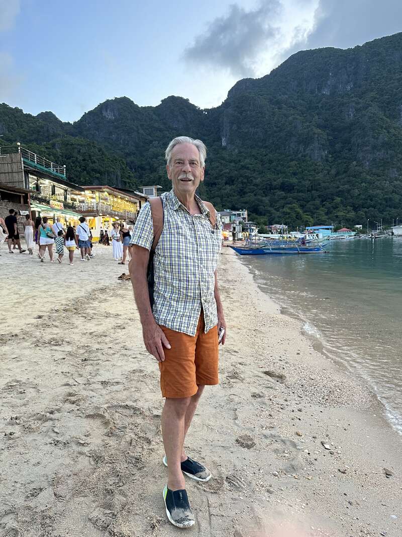 L'image représente un homme d'âge mûr debout sur une plage de sable, avec un arrière-plan montagneux et un plan d'eau à sa droite, portant une tenue décontractée et un sac à dos.