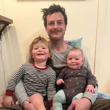 Um pai senta-se no chão com seus dois filhos pequenos, todos sorrindo alegremente. A criança mais velha usa listras, enquanto o bebê usa pijama marrom. Uma cena aconchegante.