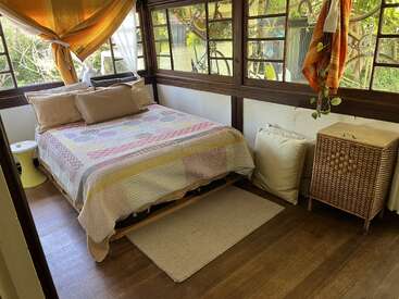 Um quarto aconchegante e ensolarado com janelas grandes, piso de madeira e uma cama bem arrumada com roupas de cama em tons pastéis. Almofadas, móveis de vime e vegetação criam um espaço acolhedor e convidativo.