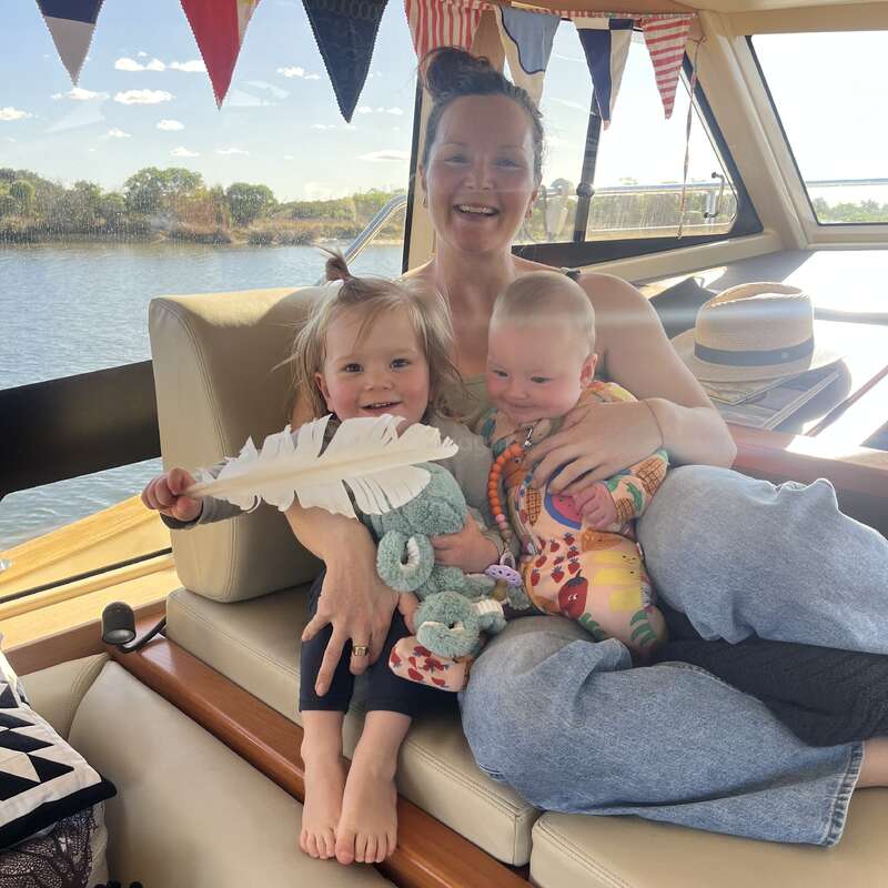 Uma mulher sorridente está sentada em um barco com duas crianças pequenas. A criança mais velha segura uma pena e um brinquedo, enquanto bandeiras festivas estão penduradas acima delas.
