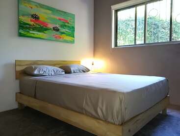 Das Bild zeigt ein ruhiges Schlafzimmer mit einem Holzbett, weißen Laken und einem lebhaften Gemälde an der Wand, das durch ein Fenster in natürliches Licht getaucht wird.