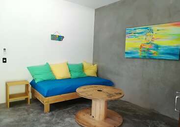 Das Bild zeigt ein gemütliches Zimmer mit einem blauen Futon, bunten Kissen, einem hölzernen Spulentisch und einem farbenfrohen Gemälde an der Wand, das ein Gefühl von Wärme und Behaglichkeit ausstrahlt.
