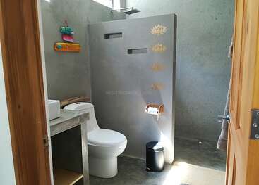 Das Bild zeigt ein Badezimmer mit einer Toilette, einem Waschbecken und einer Dusche, mit einem hölzernen Türrahmen, Betonwänden und -boden, einem Mülleimer und einem Handtuchhalter.