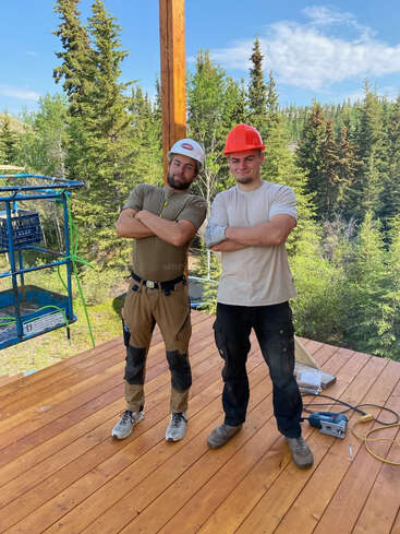 Zwei Männer stehen selbstbewusst auf einer im Bau befindlichen Holzterrasse, lächeln mit verschränkten Armen, tragen Arbeitskleidung und Schutzhelme und sind von einer üppig grünen Waldlandschaft umgeben.
