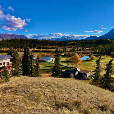 A picturesque rural scene with scattered houses, evergreen trees, a pond, open grassy fields, and majestic mountains in the background under a deep blue sky.
