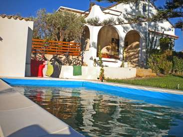 La imagen representa una serena escena de una casa blanca con arcos, una piscina y una valla, sobre un cielo azul despejado en un día soleado.