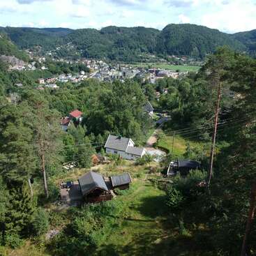 The image depicts a serene mountain town nestled among lush trees, with a cluster of houses and buildings in the foreground, set against a picturesque backdrop of rolling hills.